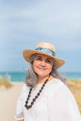 Content aged female in white dress and straw hat looking at camera while standing on coast of ocean - ADSF46144