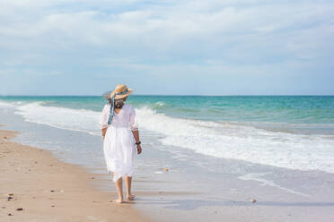 Back view of happy elderly female in white dress and straw hat walking on the beach sand - ADSF46149