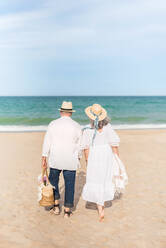 Rückenansicht eines anonymen älteren Mannes und einer Frau in weißem Kleid, die gemeinsam am Sandstrand des Ozeans spazieren gehen - ADSF46150