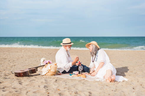 Happy senior couple on the beach opening a bottle of wine while having a picnic on the beach - ADSF46151