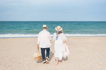 Rückenansicht eines anonymen älteren Mannes und einer Frau in weißem Kleid, die gemeinsam am Sandstrand des Ozeans spazieren gehen - ADSF46153
