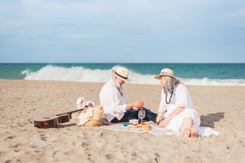 Happy man and woman in white clothes having picnic on the beach while opening a wine - ADSF46155