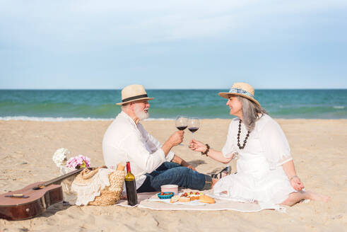 Loving aged man and woman in white dress clinking wineglasses while having picnic on sandy coast - ADSF46156