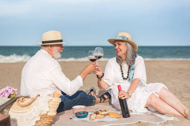 Loving aged man and woman in white dress clinking wineglasses while having picnic on sandy coast - ADSF46157
