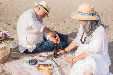 High angle of senior male in white shirt removing cork from bottle of wine while having picnic with charming lady on beach blanket - ADSF46158