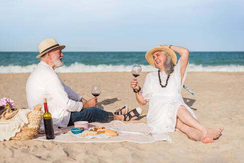 Happy couple on the beach having a picnic while holding a glass of wine - ADSF46159