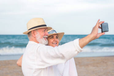 Side view positive elderly couple in white beach clothes taking selfie on smartphone while standing on sandy beach near waving sea - ADSF46163