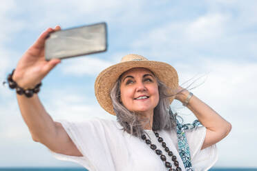 Eine zufriedene Frau mittleren Alters mit einem Strohhut macht ein Selfie an einem Sandstrand, während sie auf ihr Handy-Display blickt - ADSF46164