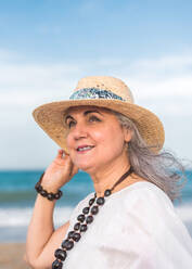 Elderly female with gray hair wearing straw hat and white dress looking away on beach in wind - ADSF46165