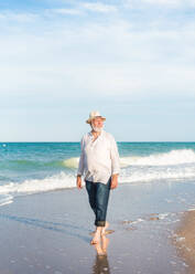 Positive middle aged barefoot male in straw hat looking away while walking on sandy beach near foamy seawater in daylight against cloudy blue sky - ADSF46168