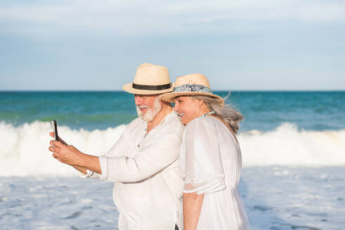 Ein Paar mittleren Alters genießt den Strand und hält Erinnerungen mit einem Selfie fest, während es das Meer auf seinem Mobiltelefon bewundert - ADSF46169