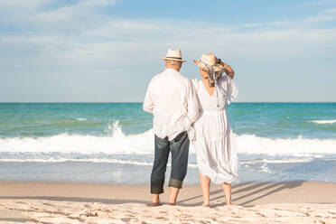 Back view of elderly man and woman in beach clothes standing watching the rolling sea on a sunny day - ADSF46173