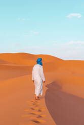 Sides view of full body of young Berber man wearing traditional Tuareg clothes walking on sand dune while looking away in Merzouga desert Morocco - ADSF46175
