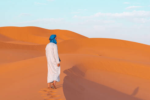 Side view of full body of young Berber man wearing traditional Tuareg clothes walking on sand dune while looking away in Merzouga desert Morocco - ADSF46176