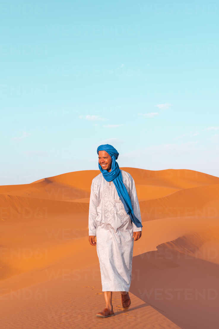 Full body of young Berber man wearing traditional Tuareg clothes walking on  sand dune while looking away in Merzouga desert Morocco stock photo