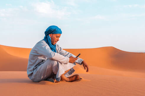 Side view of Berber man in traditional Tuareg clothes sitting on sand while browsing smartphone against blue sky in Merzouga desert Morocco - ADSF46178