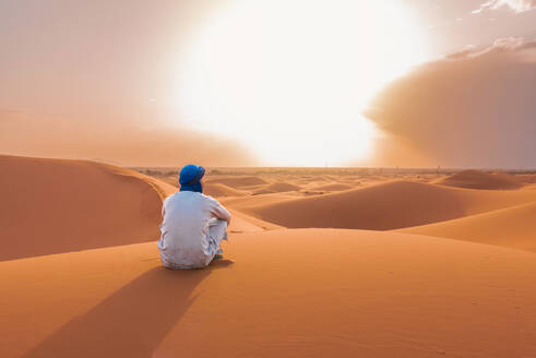 Back view of unrecognizable Berber man in traditional clothing sitting on sand dune in Merzouga desert during sunset - ADSF46179