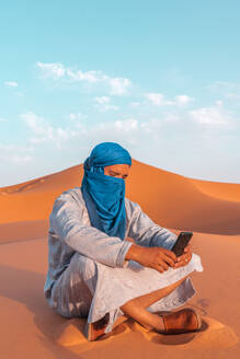 Full body of Berber man in traditional Tuareg clothes sitting on sand while browsing smartphone against blue sky in Merzouga desert Morocco - ADSF46183