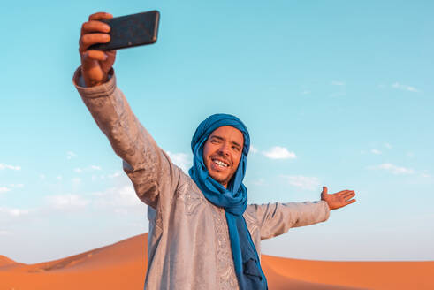 Ein junger Berber, gekleidet in traditioneller Kleidung und einem blauen Turban, macht ein Selfie mit seinem Smartphone in der marokkanischen Wüste Merzouga - ADSF46185