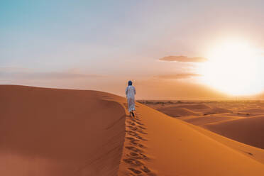 Distant back view of unrecognizable Berber man in traditional clothes walking on sand dune in Merzouga desert during sunset - ADSF46187