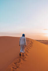 Back view of unrecognizable Berber man in traditional clothes walking on sand dune in Merzouga desert during sunset - ADSF46188