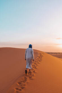 Back view of unrecognizable Berber man in traditional clothes walking on sand dune in Merzouga desert during sunset - ADSF46188