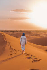 Back view of unrecognizable Berber man in traditional clothes walking on sand dune in Merzouga desert during sunset - ADSF46189