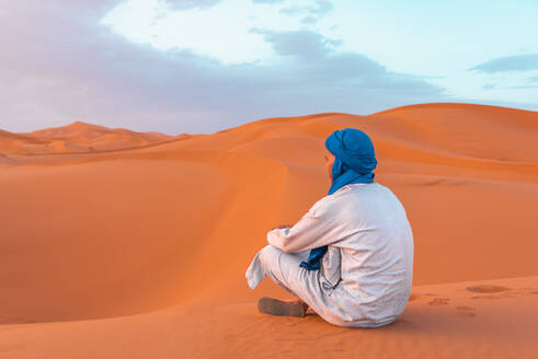 Side view of young Berber man in traditional clothing sitting on sand dune in Merzouga desert during daylight - ADSF46190