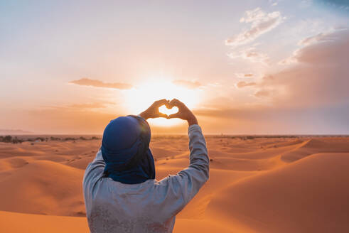 Back view of unrecognizable Berber man in traditional clothes holding hands in heart shape against sun while standing in Merzouga desert during sunset - ADSF46191