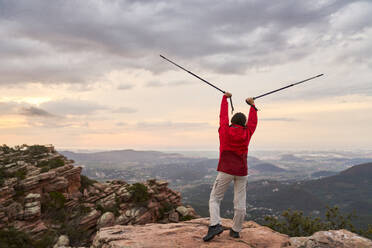 Back view of unrecognizable female traveler holding trekking poles with arms raised standing on edge of cliff El Garbi and admiring picturesque scenery of mountains - ADSF46196