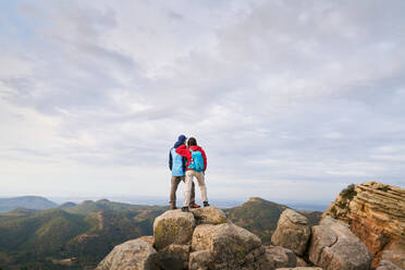 Rückenansicht einer nicht erkennbaren Frau mit Rucksack, die sich auf dem Gipfel eines Berges an ihren Mann lehnt und die majestätische Landschaft des Hochlandes in Spanien genießt - ADSF46199