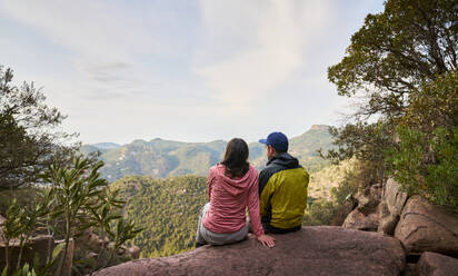 Back view of anonymous female traveler hugging boyfriend while sitting together on edge of cliff and admiring view of mountainous terrain - ADSF46202