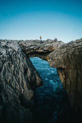 Distant male traveler standing on rocky cliff and admiring picturesque scenery of sea against blue sky in Cantabria, Spain - ADSF46210