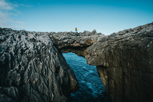 Distant male traveler standing on rocky cliff and admiring picturesque scenery of sea against blue sky in Cantabria, Spain - ADSF46211