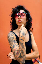 Confident young Hispanic female in sunglasses with tattooed hand finger near face looking away while standing against orange wall with bicycle in day - ADSF46216