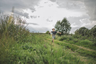 Playful boy running on field under cloudy sky - ANAF01879