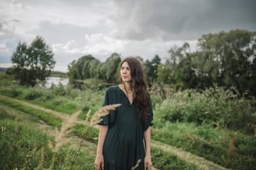 Thoughtful woman standing on field under cloudy sky - ANAF01881