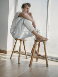 Contemplative man sitting on wooden stools at home - MFF09392