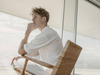 Contemplative man sitting on wooden armchair at home - MFF09394