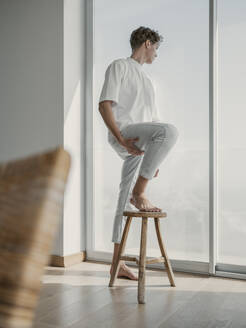 Young man with foot on stool looking through window at home - MFF09396