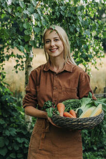 Lächelnde Frau hält Korb mit Gemüse im Garten - VSNF01255