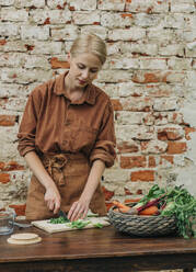 Blonde Frau schneidet Gemüse auf einem Tisch vor einer Backsteinmauer - VSNF01259