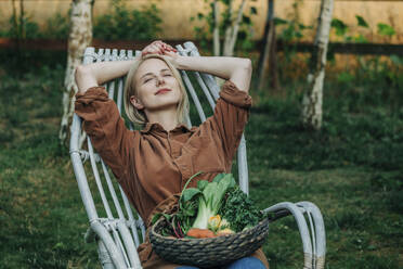 Smiling woman leaning on chair with vegetable basket in garden - VSNF01269