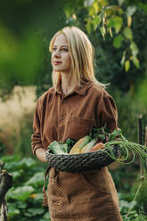 Contemplative woman holding vegetable basket in garden - VSNF01270