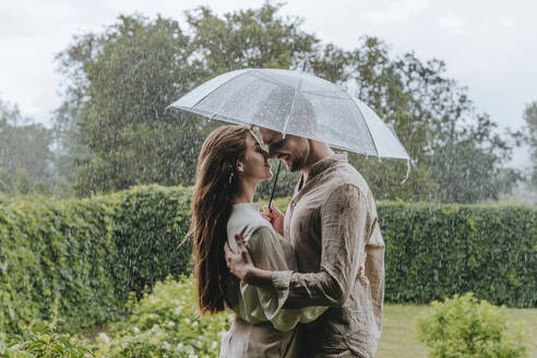 Romantic couple standing with umbrella in rain at garden - YTF00927