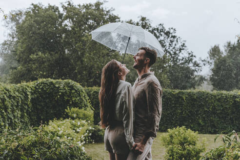 Happy couple holding umbrella and enjoying in rain at garden - YTF00928