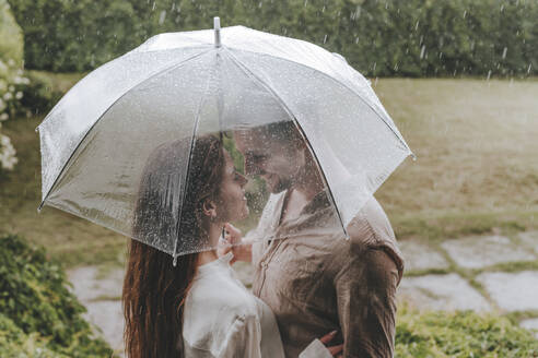 Romantic couple standing with umbrella in rain at garden - YTF00930