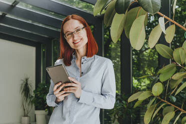 Happy businesswoman standing with tablet PC in front of window - YTF00960