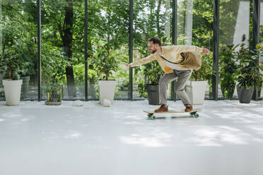 Happy businessman doing longboard skating in front of plants - YTF00965