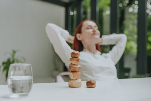 Stack of wooden pebbles on desk with businesswoman relaxing in background - YTF01006
